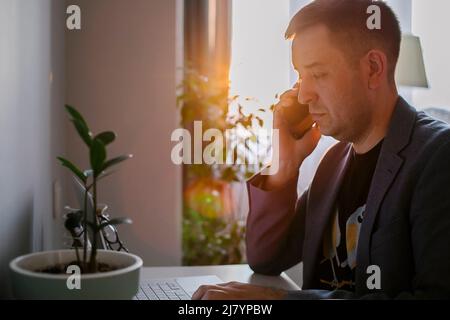 Man working remote job from home office. Cheerful freelancer working on laptop and talking on the phone Stock Photo
