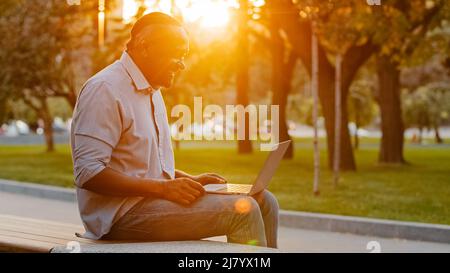 Elderly african american man sitting outdoors browsing wireless Internet on modern device, typing texting on laptop working on computer, adult Stock Photo