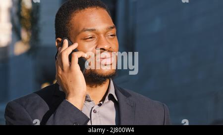 Serious focused millennial African American businessman standing outdoor talking on phone, young mixed race entrepreneur speak on cellphone, solve Stock Photo