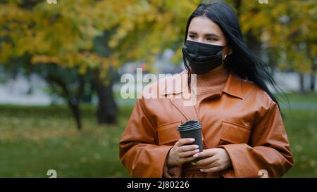 Close-up young woman stands in autumn park beautiful hispanic girl holding disposable cup of coffee wants to drink but forgets to remove medical Stock Photo