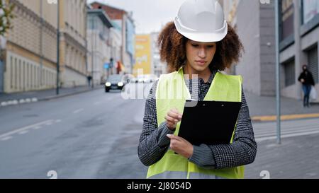 Portrait young african american beautiful female engineer wearing safety vest and hardhat standing in city near road traffic writing data Stock Photo