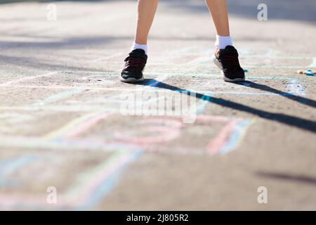 Kids play hopscotch in summer park. Healthy active outdoor game. Children playing and drawing with chalk on suburban city street. Boy and girl jump. Stock Photo