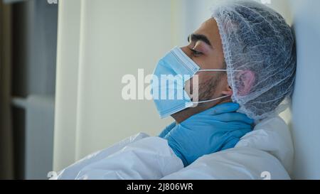Side view male doctor medical worker surgeon in protective mask sitting on floor in clinic near wall holding neck with hands feeling tired exhaustion Stock Photo