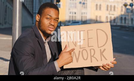 Sad mixed race young adult black man sitting outdoor with cardboard text table Need Work. Looking for job. Fired millennial upset male. Businessman Stock Photo