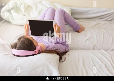 A little girl in purple pajamas, lies on a white bed on her back, holds a pink digital tablet Stock Photo