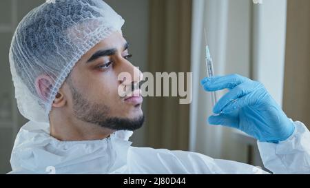 Young arabic male hispanic doctor practitioner surgeon in special medical protective uniform wears rubber gloves holds syringe with medicine vaccine Stock Photo