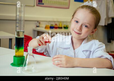 clever blonde school girl chemist in eyeglasses and white medical gown making science experiments chemistry ,mixing different chemical solutions in Stock Photo