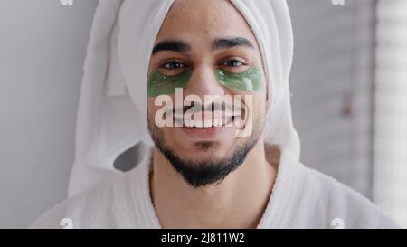 Funny portrait male face smiling indian arabian man wearing bath towel on head glue sticky hydrogel eye-patches cosmetic procedures skin care guy Stock Photo