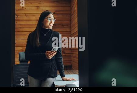 Cheerful young businesswoman looking away thoughtfully while holding a smartphone. Happy female entrepreneur sending a text message while working in a Stock Photo