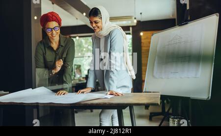 Cheerful female architects working on blueprint drawings in a modern office. Two Muslim businesswomen planning a new innovative project. Creative desi Stock Photo