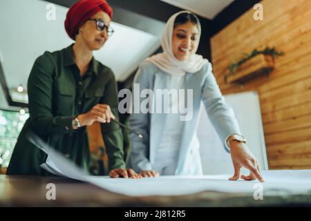 Female architects working on blueprint drawings in a modern office. Two Muslim businesswomen planning a new innovative project. Creative designers wea Stock Photo