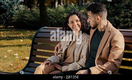 Cheerful hispanic couple cutely communicate sitting on bench in autumn city park young bearded guy hugs attractive curly girl carefree communication Stock Photo