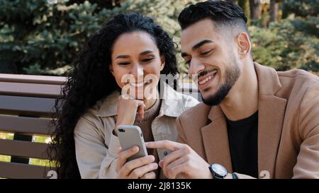 Bearded guy with attractive girl in autumn park sitting on bench cheerful couple interestedly looks at smartphone screen young man actively Stock Photo