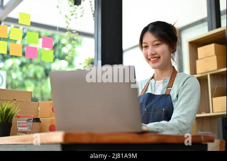Happy Asian young female fashion shop owner using laptop computer to reply message from her customer, getting a good review from customer. Small busin Stock Photo