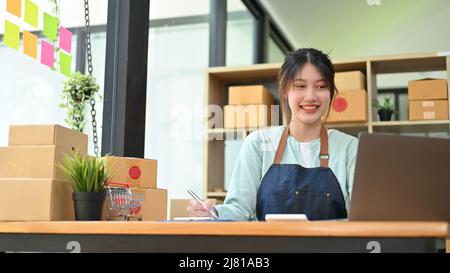 Charming Asian millennial woman checks her product stock and purchases orders from an online customer on her laptop computer. Stock Photo