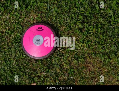 A 1 kg round pink track and field discus with a smiley face lays off center in the grass. Shot in natural sunlight with no people and copy space. Stock Photo