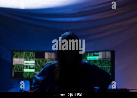 Hooded anonymous hacker by computer in the dark room at night, cyberwar concept. Stock Photo