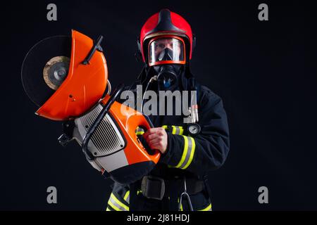 portrait strong fireman in fireproof uniform holding an ax chainsaw in his hands black background studio Stock Photo