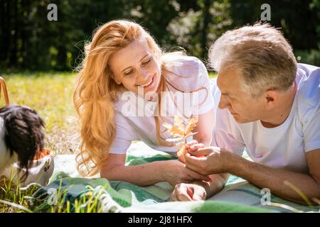 Mature couple lie with their dog in park. An elderly couple is resting in nature with dog. Close-up portrait of an elderly man and woman in white shirts and jeans. Stylish and modern grandparents. Stock Photo
