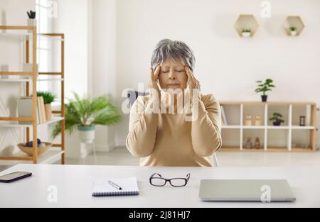 Tired senior woman who has headache after working on computer is doing head massage Stock Photo