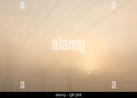 Setting sun in the mist behind three power poles with the foggy orange sky as background Stock Photo
