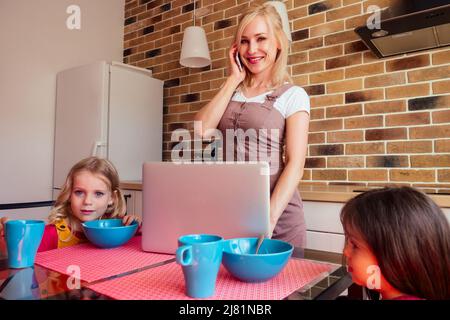 busy beautiful blonde mother with laptop stand next to two girl sisters in kitchen Stock Photo