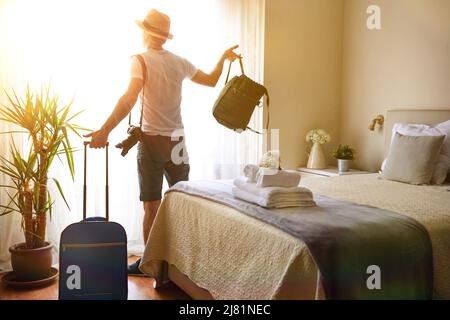 Man from behind with suitcases and handbag in a hotel room looking out the window where a ray of sunlight enters Stock Photo