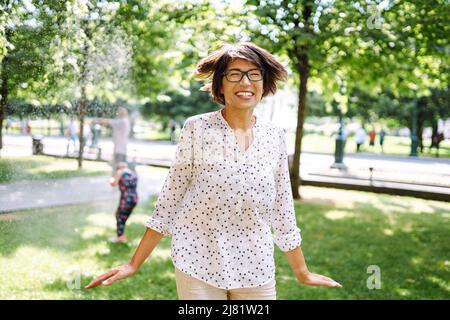 Laughing and rejoicing woman in eyeglasses jumping and playing in water stream from sprinkler fountain. Green garden in summer holiday vacation. Fresh Stock Photo