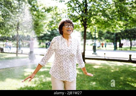 Laughing interracial woman in eyeglasses jumping and playing in water stream from sprinkler fountain. Green garden in summer holiday vacation. Freshne Stock Photo