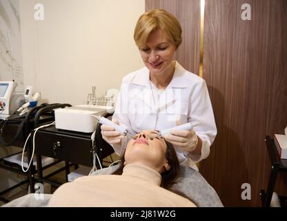 Doctor cosmetologist, aesthetician doing a professional treatment for facial muscle stimulation to regain tone on the face of a young woman. Microcurr Stock Photo