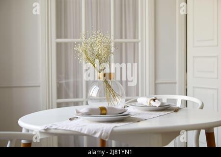 Elegant glass vase with gentle babys breath flowers and ceramic plates with rolled napkins served on round table in white dining room Stock Photo