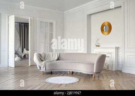 Elegant interior of spacious living room with comfortable gray velour sofa and parquet floor decorated with artificial fireplace and round gold framed Stock Photo