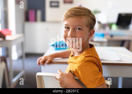 Portrait of smiling caucasian elementary schoolboy sitting at desk in classroom Stock Photo