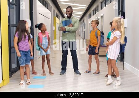African american young male teacher talking to multiracial elementary students standing in corridor Stock Photo