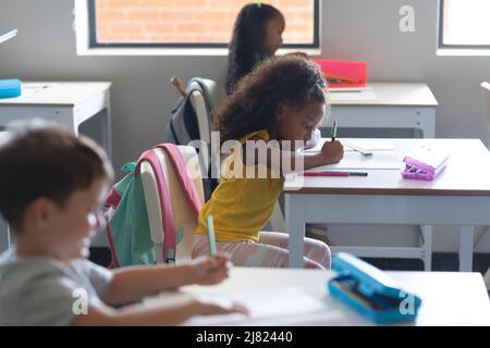 Multiracial elementary school students writing on book while studying at desk in classroom Stock Photo