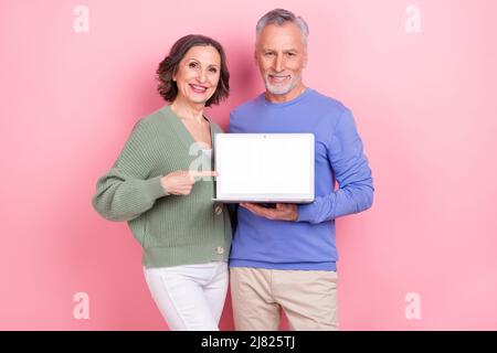 Portrait of two elderly persons direct finger empty space netbook display isolated on pink color background Stock Photo