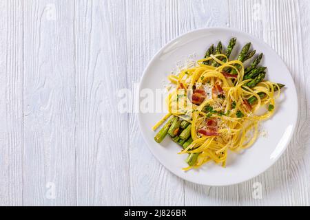 roasted asparagus spears with spaghetti sprinkled with shredded cheese, fried bacon pieces and parsley on white plate on white textured wood table, fl Stock Photo