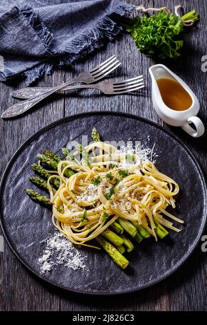 close-up of roasted asparagus spears with spaghetti sprinkled with shredded pecorino cheese and parsley on black plate on dark wood table, vertical vi Stock Photo