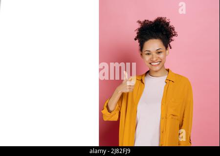 Positive pretty, curly african american girl, in casual wear, shows thumb up gesture, stand near blank white space for advertising or presentation, on pink background, looks at camera, smiles. Mock-up Stock Photo