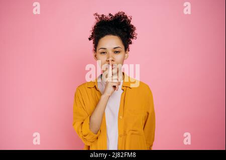 Portrait of a pretty, curly haired african american girl, showing shh sign with finger near lips, silence gesture, secret concept, standing on isolated pink background, looking at the camera Stock Photo