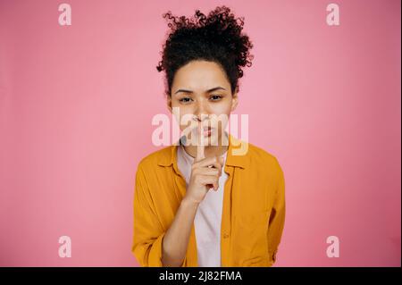 Silence gesture, secret concept. Lovely african american girl in casual wear, showing shh sign with finger near lips, silence gesture, standing on isolated pink background, looking at the camera Stock Photo