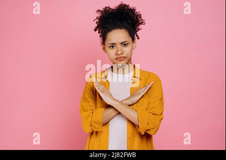 Strict curly haired african american girl in casual clothes, saying no, crossed hands in stop gesture, expressing dissatisfaction, standing on isolated pink color background, looking at camera Stock Photo