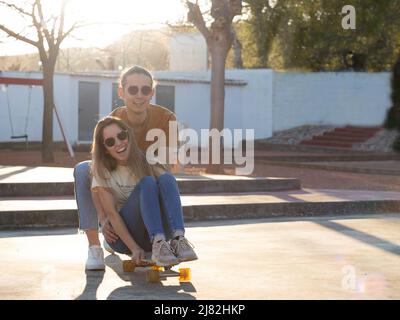 Summer love between a couple of teenagers having a good time on a skate board in the city park at sunset time Stock Photo