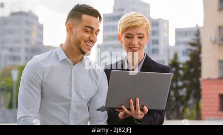 Serious diverse multiracial couple colleagues arabic hispanic man and caucasian woman standing near business center at street looking at computer Stock Photo