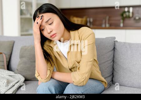 Stressed young asian woman touching head and feels dizzy, suffering from headache and migraine pain, sits on the sofa at home, tired and exhausted lady feels bad Stock Photo