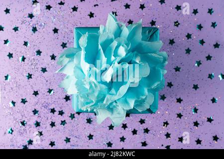 A gift wrapped in blue eco kraft paper with a lush bow on a lilac background with small confetti in the form of stars.The holiday, surprise, birthday. Stock Photo