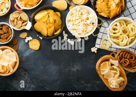 Unhealthy food. Snacks. All classic potato snacks with peanuts, popcorn and onion rings and salted pretzels in bowl plates on black concrete backgroun Stock Photo
