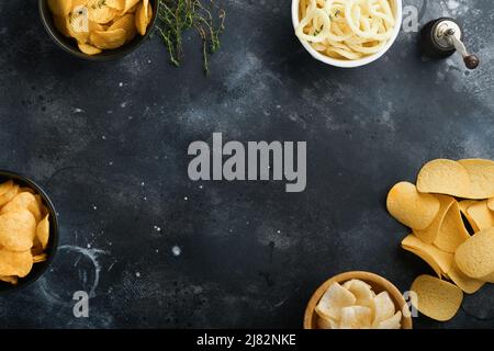 Unhealthy food. Snacks. All classic potato snacks with peanuts, popcorn and onion rings and salted pretzels in bowl plates on black concrete backgroun Stock Photo