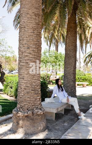 full length view of woman sitting on bench in park under giant palms Stock Photo