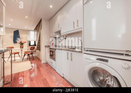 Long apartment kitchen with white cabinets, pink granite countertop, round white dining table, and reddish wood flooring Stock Photo